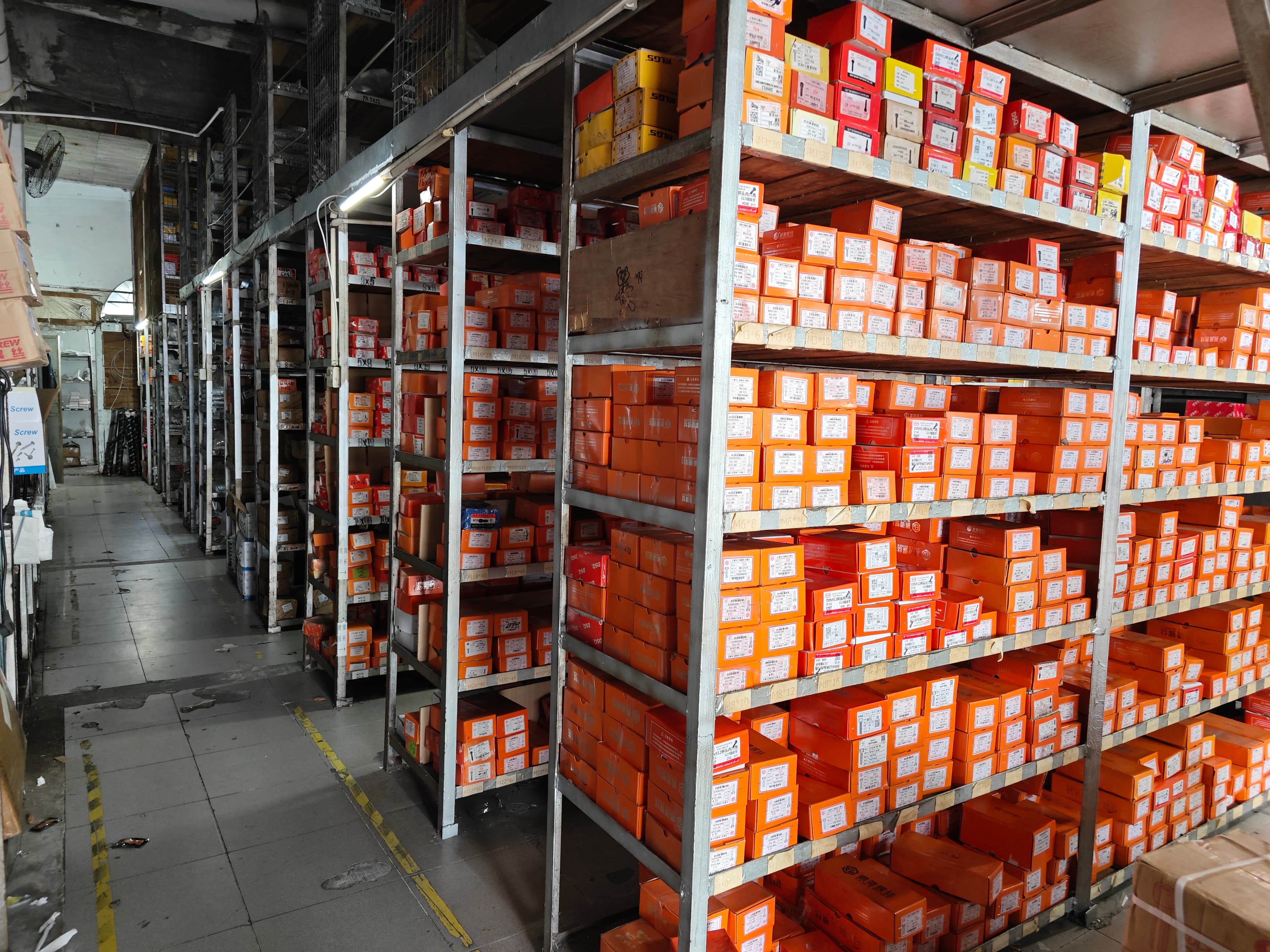 An aisle beside tall shelving filled with labelled orange and red fastener boxes.